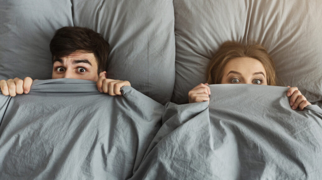 Surprised couple hiding under grey sheets in bed at home, covering half face, looking at camera, top view