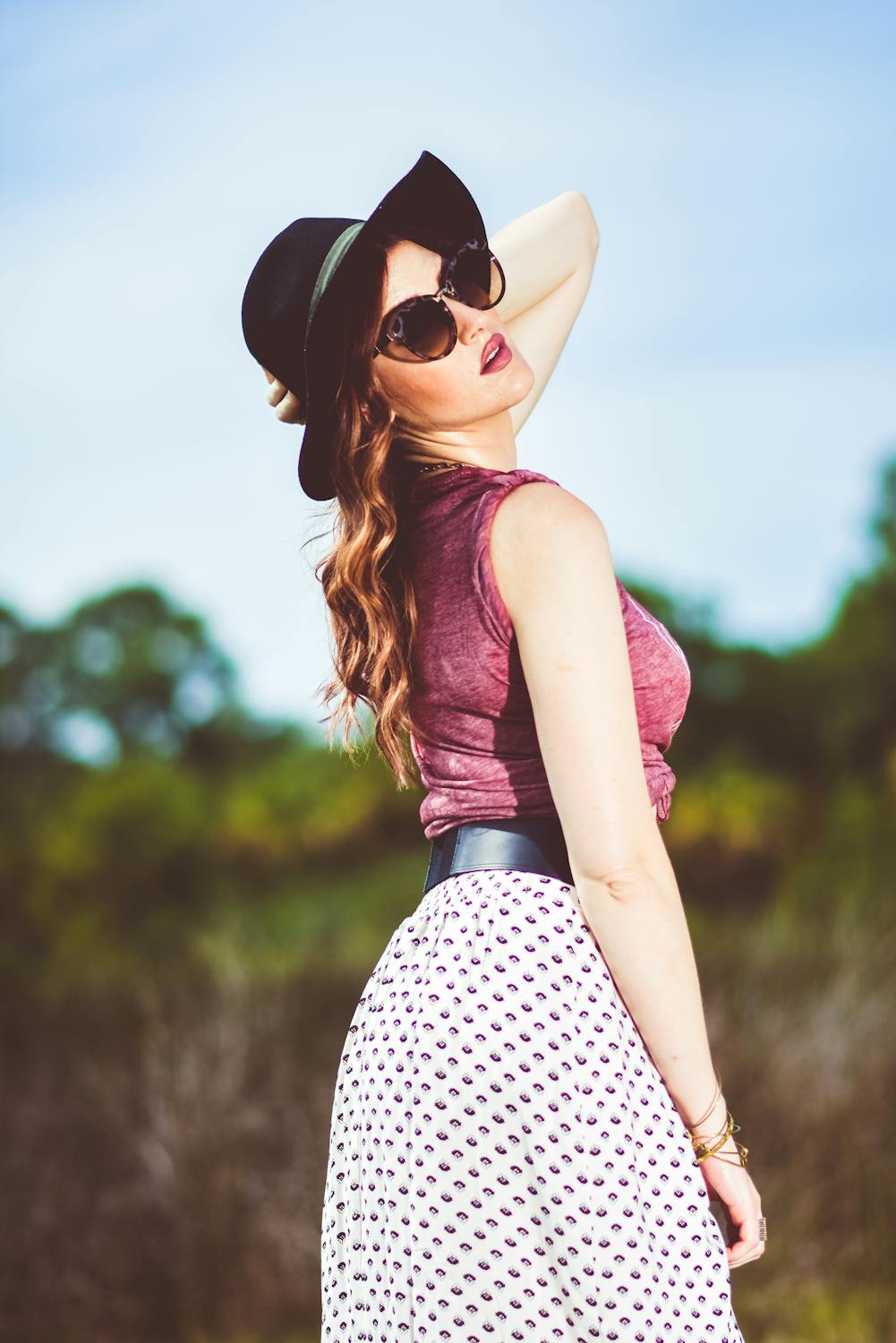 A stylish woman wearing sunglasses, a black hat, a maroon top, and a patterned skirt poses outdoors under a bright blue sky, embodying a chic and effortless vacation look.