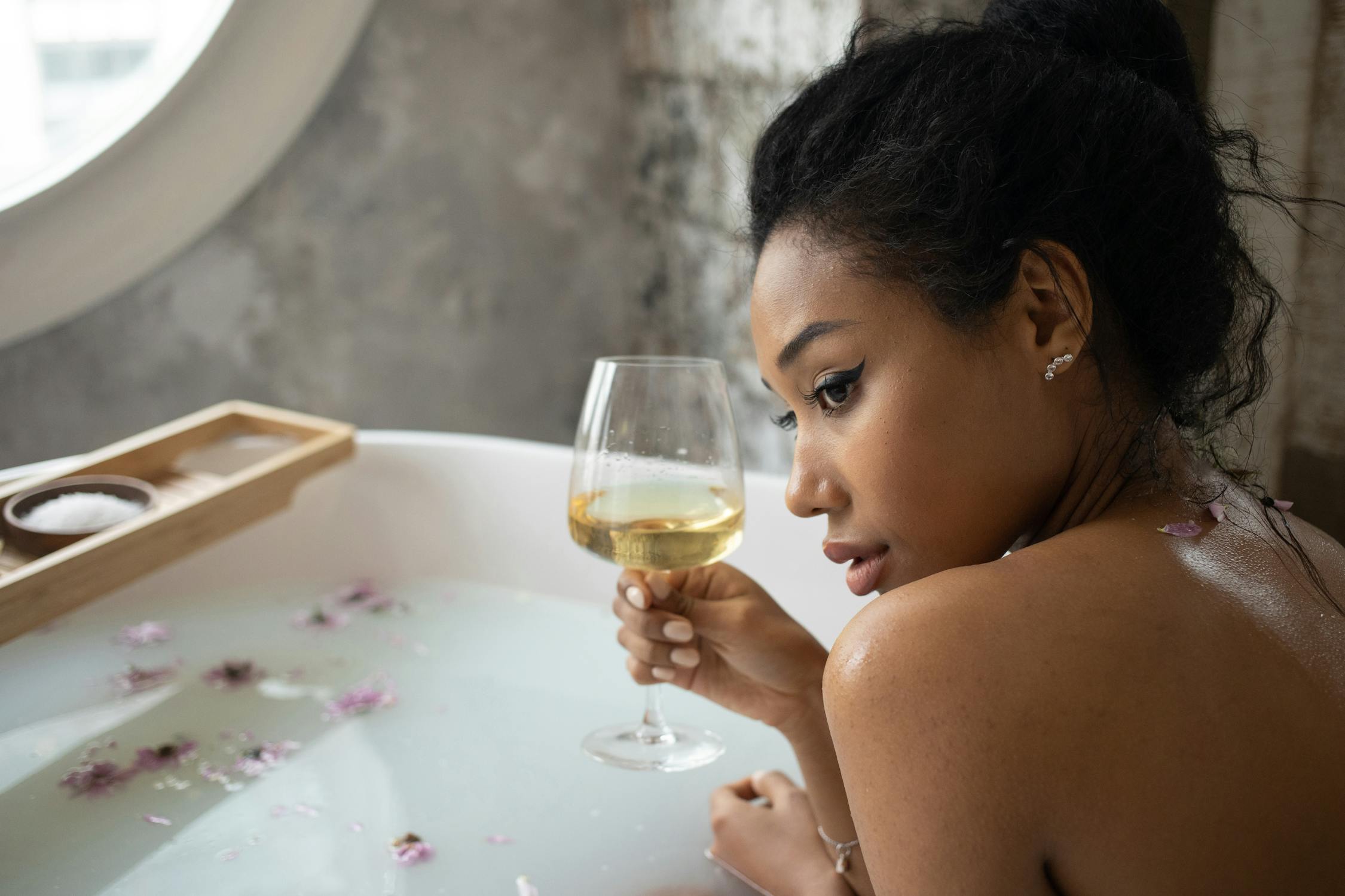 A woman with dark hair tied in a messy bun relaxes in a bathtub filled with milky water and flower petals while holding a glass of white wine. A wooden bath tray with bath salts rests on the tub’s edge.