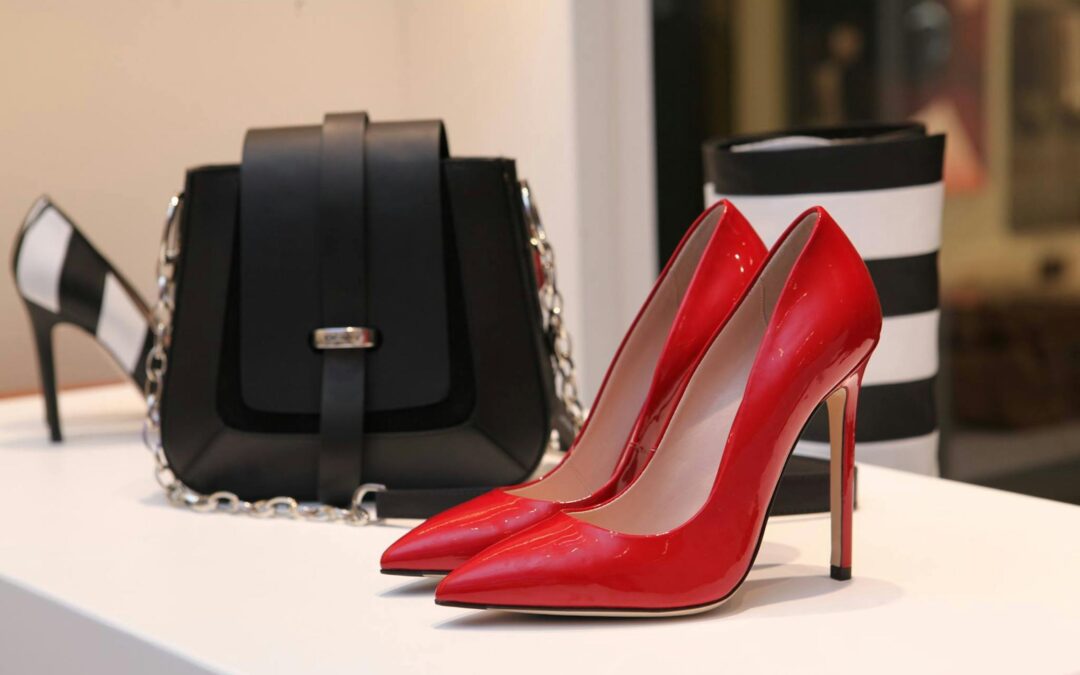 A pair of glossy red high heels displayed on a white surface, accompanied by a black designer handbag with a silver chain strap and a black-and-white striped accessory in the background.