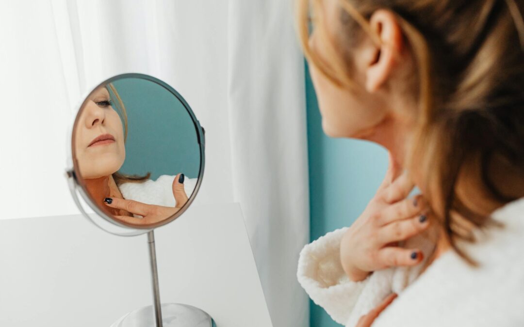 A woman in a white robe examines her skin in a round mirror while gently touching her neck, reflecting a self-care and skincare routine.